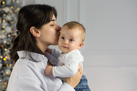 A young beautiful mother kisses her little child while standing near a Christmas treeの写真素材