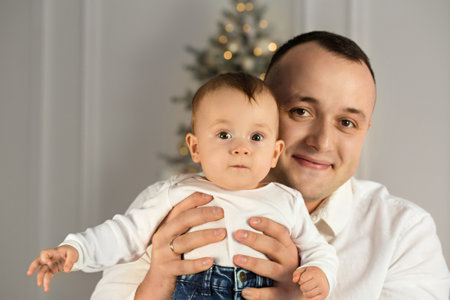 Portrait of a young happy dad with a baby in his arms, smiling near the Christmas treeの写真素材