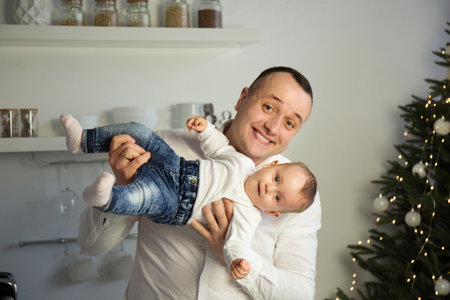 Portrait of a young happy dad with a baby in his arms, smiling near the Christmas treeの写真素材