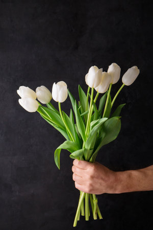 A male hand holds a bouquet of white tulips on a black backgroundの写真素材