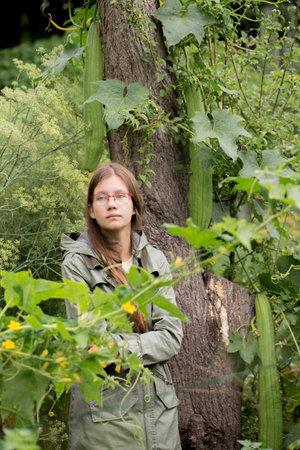 A teenage girl in a jacket stands near a loofah plant in the garden. She is wearing a green jacket...