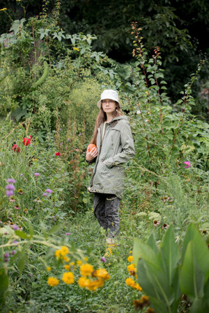 Girl wearing a jacket and a white hat walks through the garden with flowers holding a pumpkinの写真素材