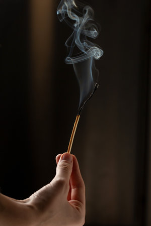 A woman's hand holds a long match that is extinguished and smoking on a dark backgroundの写真素材