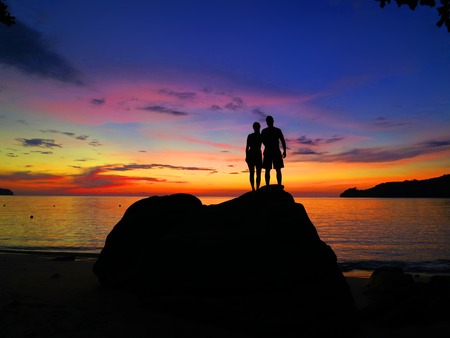 romantic couple standing on rocks by sea on purple dramatic sunset backgroundの写真素材