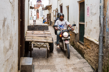 Zanzibar, Tanzania - May 05 2018: Two local men (African, Arabian, or Indian) driving a motorcycle through narrow streets of Stone Town, old colonial center of Zanzibar City, Unguja island, Tanzania.のeditorial素材