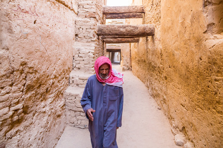 Dakhla Oasis, Egypt - Nov 23, 2018: old Arabian Egyptian man walking dressed in blue national male dress thawb, medieval street through ancient islamic town in Al Qasr built of mud bricks (motion).のeditorial素材