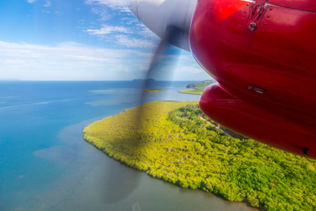 Air travel in Fiji, Melanesia, Oceania. View of a green remote tropical island from a window of a small turboprop propeller red engine airplane flying over blue azure turquoise lagoon, near Viti Levu.の写真素材