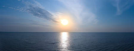 Panorama of sunrise on the sea. Small waves on the water and a cloudy sky with orange sun. Quiet morning rest on the beachの写真素材