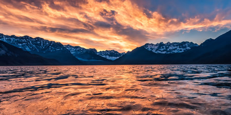 Wide angle majestic view of rocky mountains covered with snow near a lake under a cloudy sunset sky.の素材