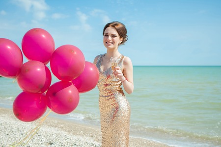 celebration and lifestyle concept - beautiful happy woman in gold dress with red balloons, glass champagne on the seaの写真素材