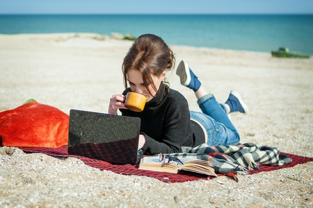 Enjoying her working day. Attractive young woman lying on a comfortable blanket on the beach. Working or studying with laptop and book.の写真素材