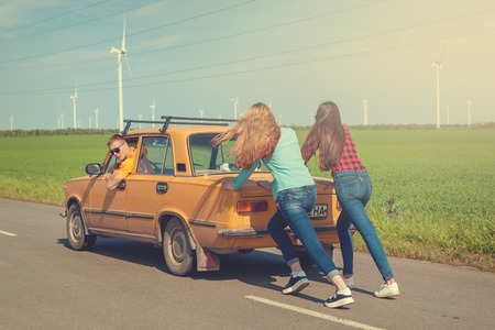 Young hipster friends on road trip on a summers day. Engine break down.Two girls pushing a vintage car while man is emboldening their.Travel, adventure, unforeseenteamwork, funny concept. Retro tonedの写真素材