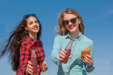 Friends and juice or smoothie. Two young cheerful girls with juice and buns smiling  on background of blue skyの写真素材