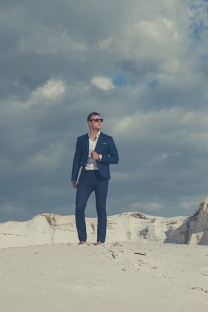 Beautiful young man in suit stands on top of hill sand  while in desert with laptop enjoying nature and the sun. Peaceful place to work and leisure. Achieving successの写真素材