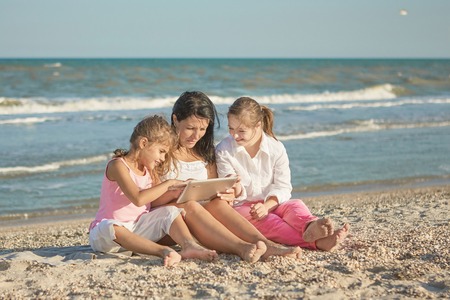 Happy family. Loving mother and two daughter on the beach play with the tablet. One daughter with a disability with Down syndrome. Positive human emotions, feelings.の写真素材