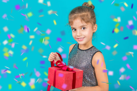 Beautiful little happy girl with a gift in a red box with ribbons on a blue background and confettiの写真素材