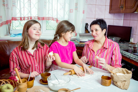 Mom teaching her two children cooking on the kitchen. Parent together with  youngest daughter and daughter with Down syndrome making pie and cookies. Family at home lifestyle photo.の素材 [FY31099276576]