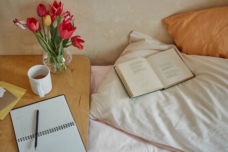 Wooden table with paper notebook, book, cup of tea and spring flowers next to bed. Good morning concept.の写真素材