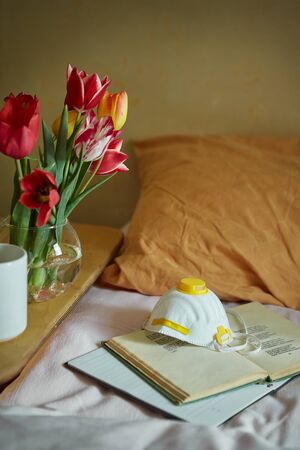 Wooden table with paper notebook, book, medical mask, cup of tea and spring flowers next to bed. Good morning quarantine concept.の写真素材