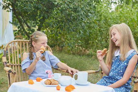 Two little girls are sitting in armchairs, having tea, in summer garden. Tea drinking, fun.の写真素材