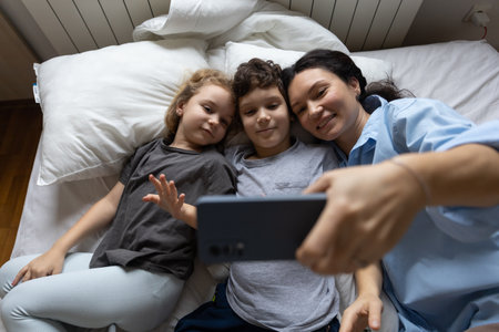 Happy caucasian mother and 2 kids have online video call with father or grandmother mobile phone when lying on bed at home. the concept of modern communication.lifestyle. top view High quality photoの写真素材