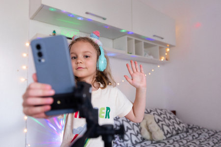 A little girl blogger wearing headphones records a video or story on her phone in her room against a background of color lights and flashlights. The content creator records vlog.の写真素材