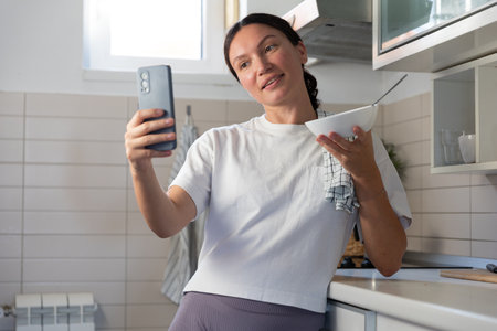 A housewife girl stands in the kitchen with a phone and a plate in her hands and blogs about cooking and how to prepare food. online blog.の写真素材