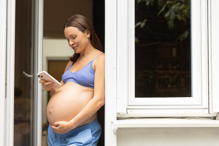 A pregnant woman is standing with a phone with a big belly in her house, using a mobile application. Space for textの写真素材