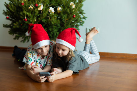 2 children in red hats on the background of Christmas tree using phone, new year mood at homeの写真素材