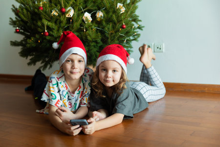 2 children in red hats on the background of Christmas tree using phone, new year mood at homeの写真素材