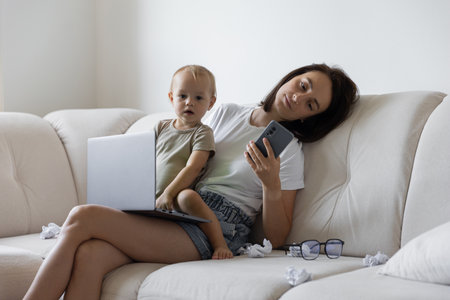 Young girl sitting on the couch and trying to work next to the child. Procrastination and deadline at workの写真素材