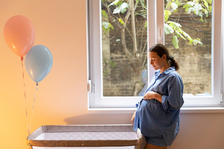 Young mom standing by crib and stroking her belly, concept of expecting baby and first pregnancyの写真素材