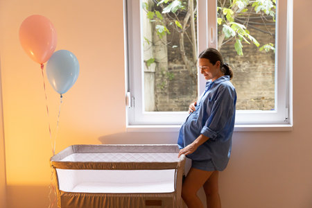 Young mom standing by crib and stroking her belly, concept of expecting baby and first pregnancyの写真素材