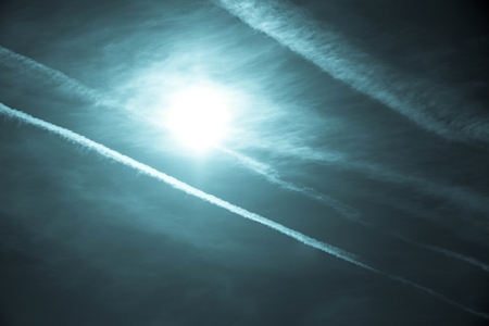 the bright white sun and traces from white exhausts of jet planes in the dark turquoise skyの写真素材