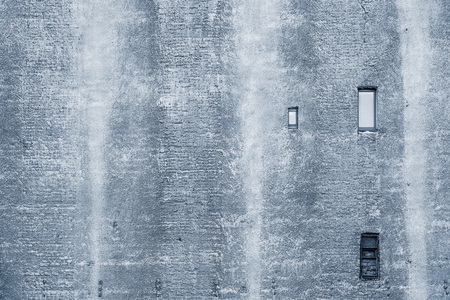 big brick wall of the old building of silvery color with the damaged plaster and with small windowsの写真素材