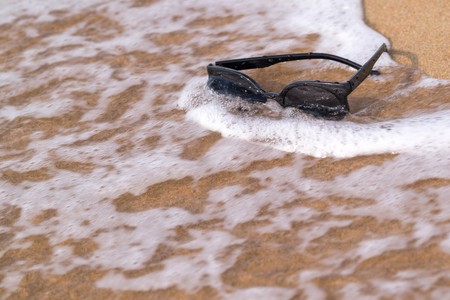 one sunglasses of black color are located separately a closeup on the sandy coast with a sea waveの写真素材