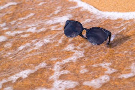 one sunglasses of black color are located separately a closeup on the sandy coast with a sea waveの写真素材