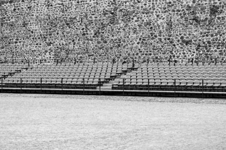 rows of seats for the audience in the foreground outdoors and against the background of an ancient stone wall of monochrome toneの写真素材