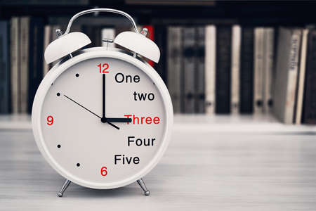 The desktop clock alarm clock is located closeup in the foreground on a blurve shelf background with the books and show a time of 3 three or 15 fifteen hoursの写真素材