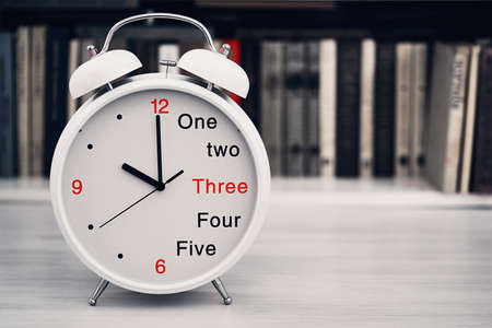 The desktop clock alarm clock is located closeup in the foreground on a blurve shelf background with the books and show a time of 10 ten or 22 twenty-two hoursの写真素材