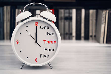 The desktop clock alarm clock is located closeup in the foreground on a blurve shelf background with the books and show a time of 12 twelve or 24 twenty-four hoursの写真素材