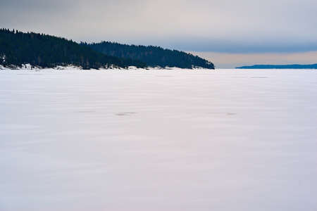 winter landscape with white snow on a large lake or in a clear field and with forest on a hilly horizonの写真素材