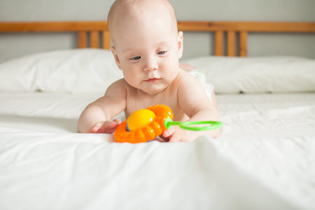 A cute baby lies on a white sheet and plays cheerfully with a bright rattle, examines it, holds it.