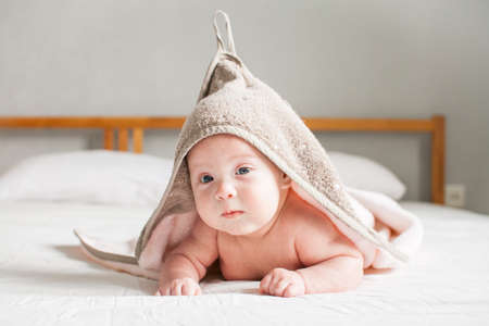 Bath time. Cute infant baby looking at camera wrapped in towel after bathing in the bath on white bed. Copyspaceの写真素材