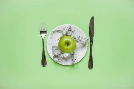 Diet and healthy eating concept. Top view of weightloss. Green apple and measuring tape on a plate, knife with a fork. Green backgroundの写真素材