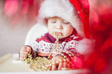 A little baby girl in a New Years hat of Santa Claus examines and plays with New Years decorations. Merry Christmas and Happy New Year greetings.の写真素材