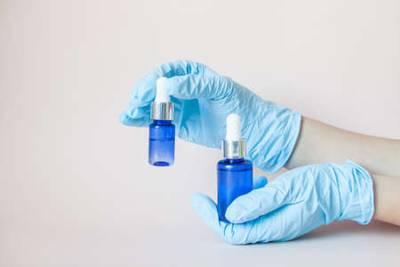Bottles with serum for the face in his hands in protective medical rubber gloves on a light background. Beauty industry.の写真素材