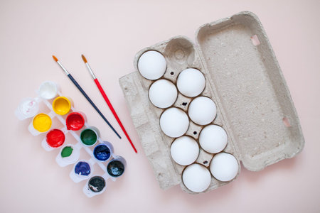 Bright paints and two brushes on a light pink background. Egg cassette. Preparing to paint eggs for...