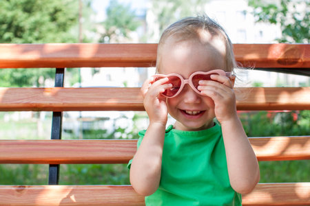 Little girl in a green T-shirt and pink heart-shaped sunglasses walks in the park in summer. High...