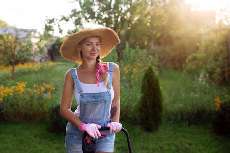 Women gardener. A sexy girl in a hat mows the grass near the house with a lawnmower. Landscape design and lawn careの写真素材